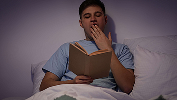 Junger müder Mann sitzt im Halbdunkeln im Bett und liest.