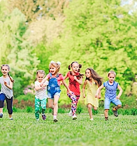 Eine Gruppe kleiner Kinder rennt über eine grüne Wiese. Sie haben Spaß zusammen und lachen ausgelassen. 