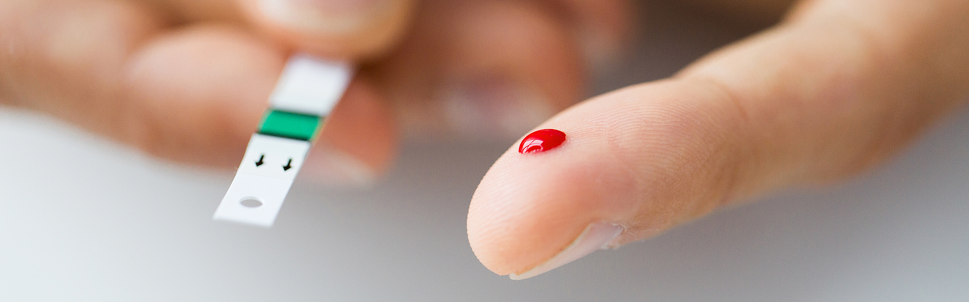 Es sind zwei Hände zu sehen. Die eine hält den Teststreifen. Bei der anderen wurde in den Zeigefinger gestochen. Es ist ein Blutstropfen zu sehen. Jemand misst seinen Blutzuckerwert.