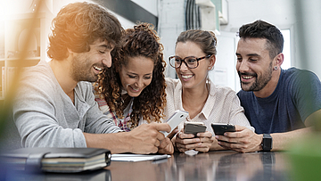 Zwei glückliche Paare sitzen am Tisch und schauen gemeinsam in ihre Handys.