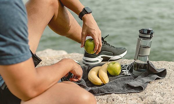 Ein Sportler sitzt am Ufer. Er ißt einen Apfel. Vor ihm liegen noch Bananen.