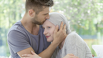 Paar sitzt zusammen auf dem Sofa. Mann küsst der kranken Frau auf die Stirn.