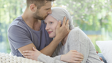 Paar sitzt zusammen auf dem Sofa. Mann küsst der kranken Frau auf die Stirn.