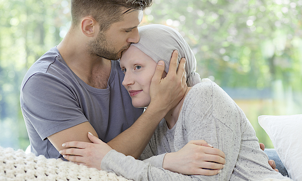 Paar sitzt zusammen auf dem Sofa. Mann küsst der kranken Frau auf die Stirn.