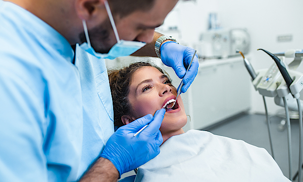 Eine Frau liegt auf einem Behandlungsstuhl beim Zahnarzt. Der Arzt untersucht ihr Zähne mit seinen Geräten. Sie schaut verunsichert.