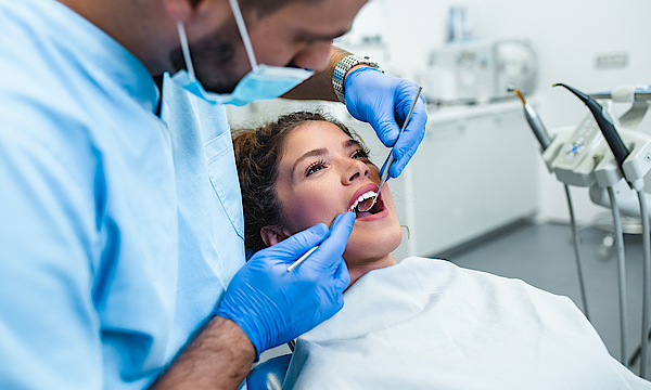 Eine Frau liegt auf einem Behandlungsstuhl beim Zahnarzt. Der Arzt untersucht ihr Zähne mit seinen Geräten. Sie schaut verunsichert.