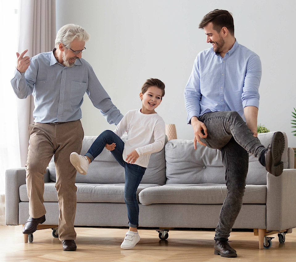 Opa, Kind und Vater machen gemeinsam Gymnastik vor einem grauen Sofa.