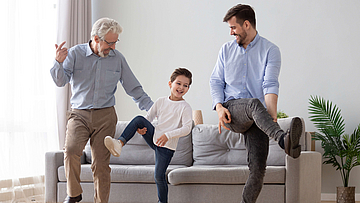Opa, Kind und Vater machen gemeinsam Gymnastik vor einem grauen Sofa.