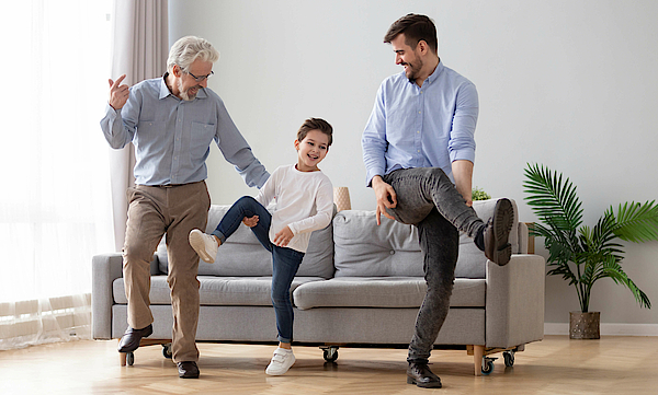 Opa, Kind und Vater machen gemeinsam Gymnastik vor einem grauen Sofa.