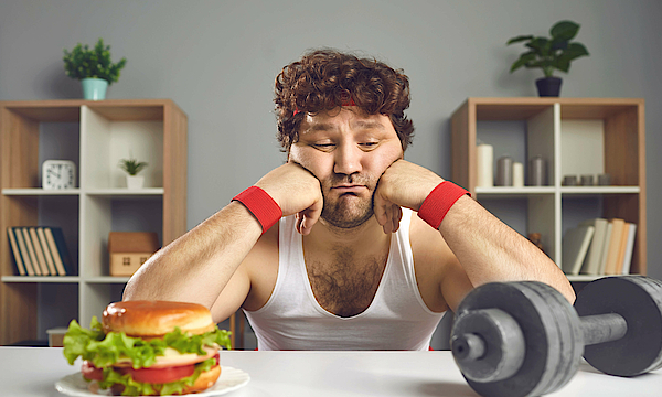 Ein junger Mann mit Sportkleidung sitzt an einem Tisch. Den Dopf hat er auf seine Fäuste gestützt. Er blickt sehnsüchtig auf einen leckeren Hamburger. Daneben liegt eine Hantel.