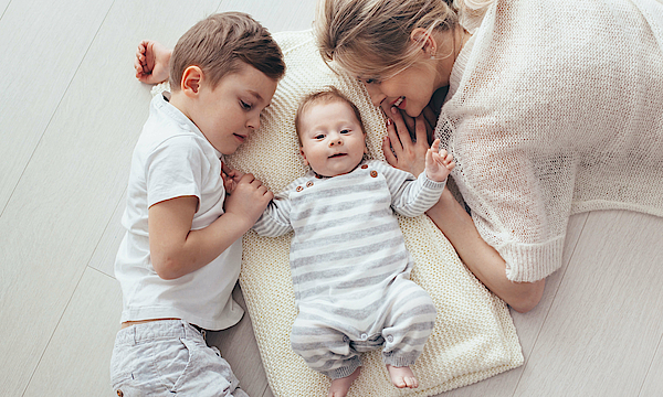 Eine Mutter liegt mit ihrem Baby und den großen Bruder auf einer Decke. Die drei kuscheln aneinander. 