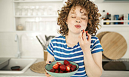 Eine junge Frau steht mit geschlossenen Augen in einer Küche. Dabei hält sie eine Schale Erdbeeren in der Hand. Sie hat soeben in eine Erdbeere gebissen und genießt den Geschmack.