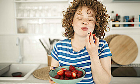 Eine junge Frau steht mit geschlossenen Augen in einer Küche. Dabei hält sie eine Schale Erdbeeren in der Hand. Sie hat soeben in eine Erdbeere gebissen und genießt den Geschmack.