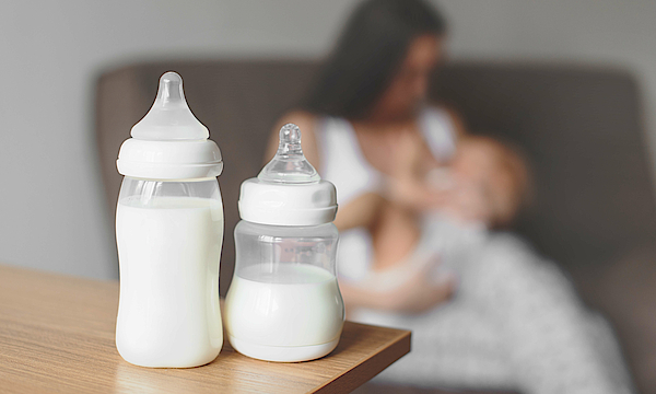 Zu sehen sind zwei Nuckelflaschen mit Milch gefüllt und im Hintergrund eine Frau die ihr Kind stillt.