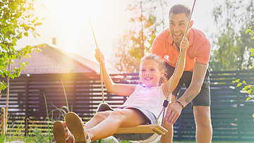 Ein Mädchen sitzt im Garten auf einer Schaukel. Sie wird von einem Mann angeschuckt beide lachen.