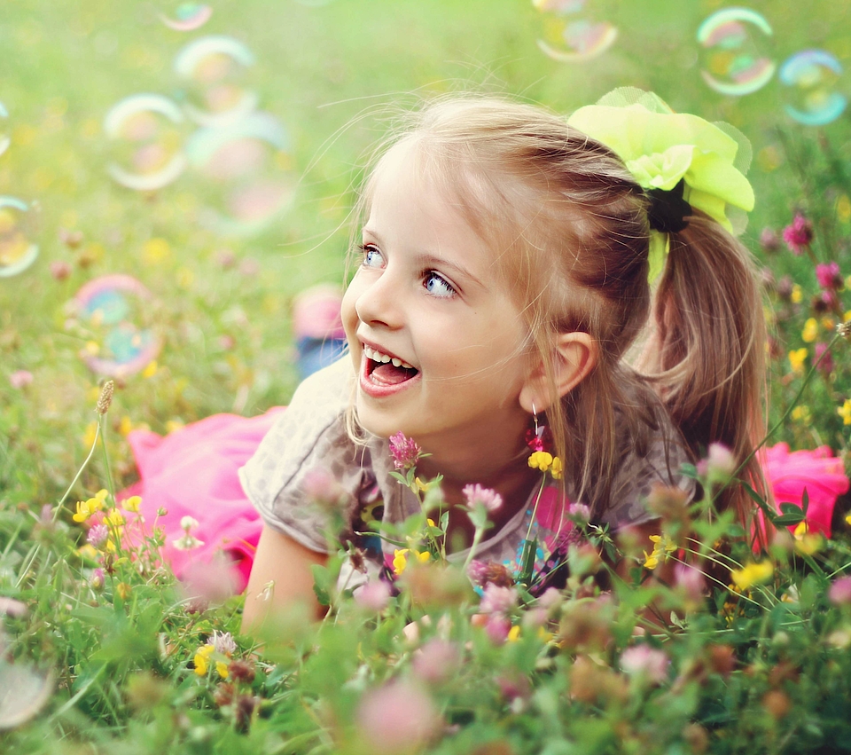 Ein kleines Mädchen mit blonden Haaren und Zöpfen liegt auf einer grünen Wiese. Um sie herum fliegen Seifenblasen. Es ist Sommer. Sie lacht glücklich und schaut den Seifenblasen nach.