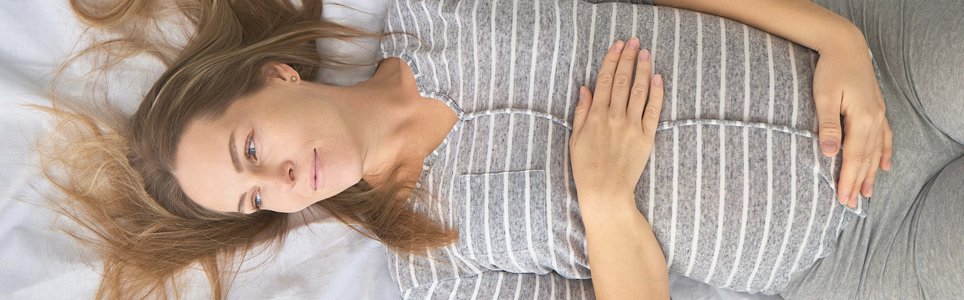 Eine schwangere blonde Frau liegt auf einem Bett. Mit beiden Händen umfasst sie ihren Bauch. Sie schaut nachdenklich.