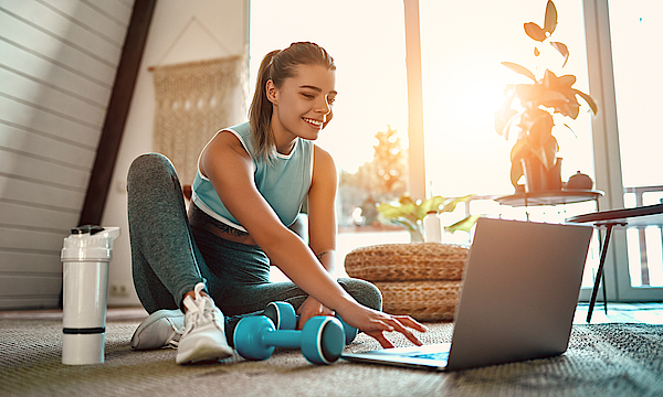 Eine sportliche Frau in Sportbekleidung sitzt auf dem Boden mit einer Hantel und einer Flasche Wasser und benutzt zu Hause im Wohnzimmer einen Laptop. 