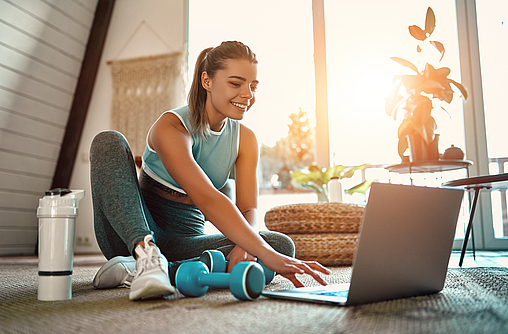 Eine sportliche Frau in Sportbekleidung sitzt auf dem Boden mit einer Hantel und einer Flasche Wasser und benutzt zu Hause im Wohnzimmer einen Laptop. 