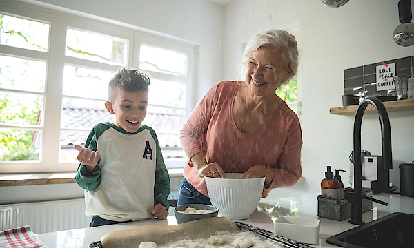 Enkel steht mit der Oma in der Küche, sie backen gemeinsam. Das Backblech ist schon fast voll.