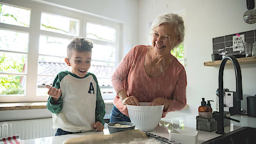 Enkel steht mit der Oma in der Küche, sie backen gemeinsam. Das Backblech ist schon fast voll.