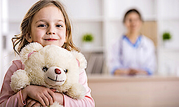 Kleines Mädchen schaut mit einem Teddy in die Kamera, Eine Frau steht im Hintergrund.