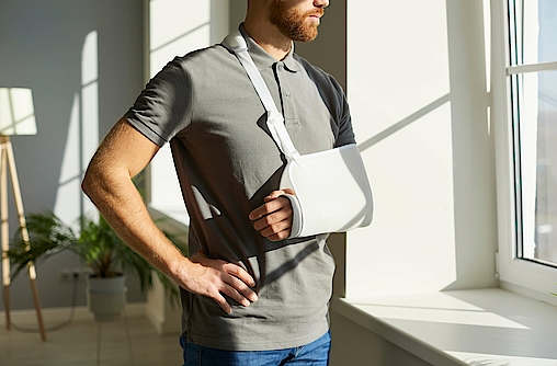 Orthopädie-Notfall. Verletzten Mann, der während der Rehabilitation zu Hause mit medizinischer Stütze und Sling Bandage Wegfahrsperre auf seinem Arm getragen. Der junge Mann mit gebrochenem Arm sieht sorgfältig aus dem Fenster.