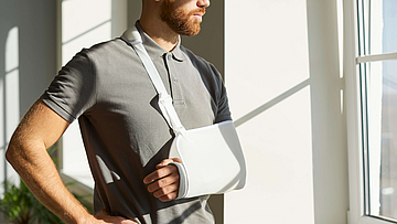 Orthopädie-Notfall. Verletzten Mann, der während der Rehabilitation zu Hause mit medizinischer Stütze und Sling Bandage Wegfahrsperre auf seinem Arm getragen. Der junge Mann mit gebrochenem Arm sieht sorgfältig aus dem Fenster.