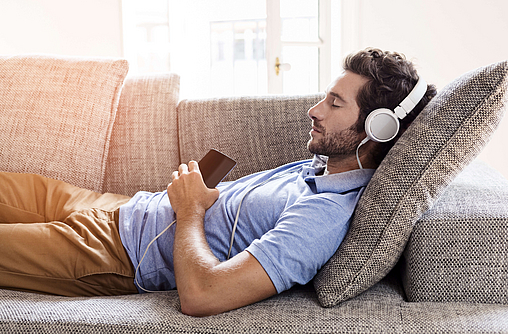 Zu sehen ist ein Mann der mit Kopfhörern auf dem Sofa liegt