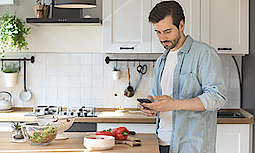 Ein Mann steht in der Küche und schaut während der Zubereitung des Essens auf sein Handy.
