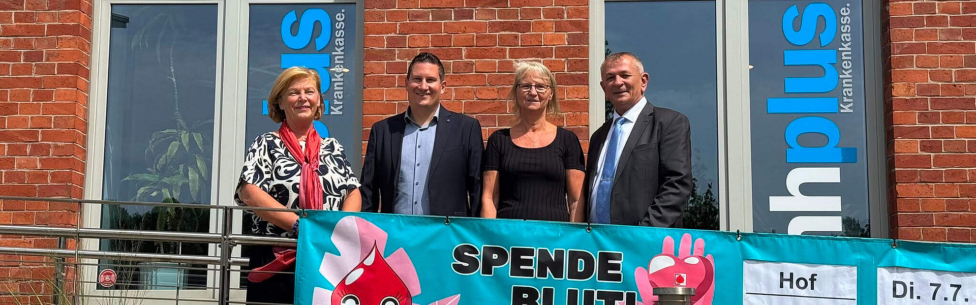 mhplus-Geschäftsstellenleiter Daniel Strötz steht vor der mhplus-Geschäftstelle in Hof zusammen mit einer Blutspenderin, der Bürgermeisterin und dem stellvertretenden Landrat vor einem Plakat zur Blutspendeaktion.