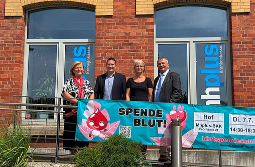 mhplus-Geschäftsstellenleiter Daniel Strötz steht vor der mhplus-Geschäftstelle in Hof zusammen mit einer Blutspenderin, der Bürgermeisterin und dem stellvertretenden Landrat vor einem Plakat zur Blutspendeaktion.