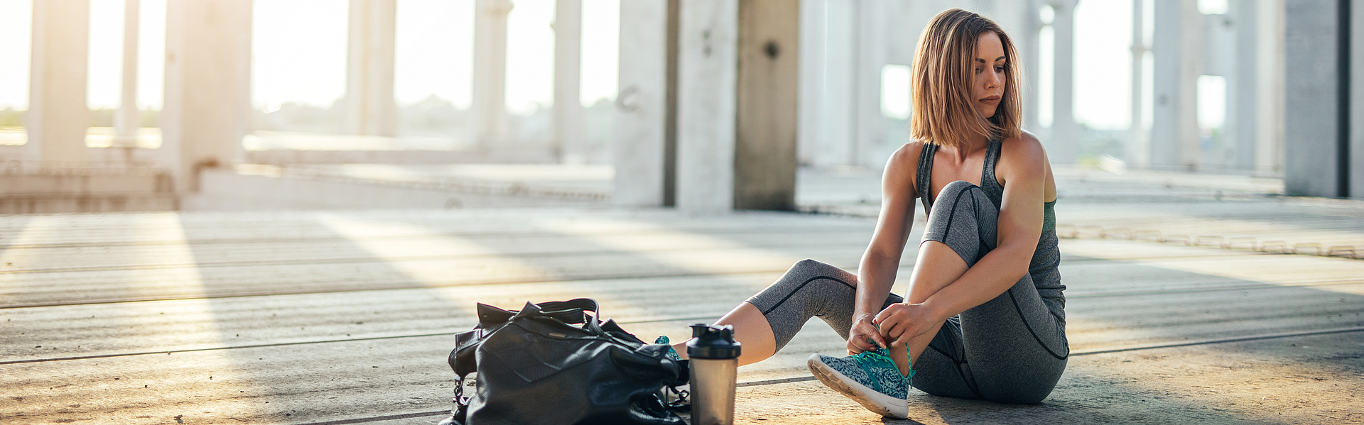 Frau zieht im Freien ihre Laufschuhe an. Die geöffnete Sporttasche und eine Trinkflasche sind auf dem Boden.