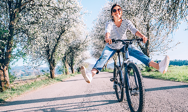 Frau fährt mit dem Fahrrad und hat die Beine weit von den Pedalen weggestreckt. Sie fährt auf einem Kiesweg zwischen blühenden Bäumen.