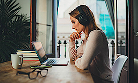 Eine junge Frau sitzt an einem Tisch und betrachtet nachdenklich ihren Laptop. Sie trägt ein schwarzes, kabelloses Headset. Im Hintergrund ist eine Pflanze und eine geöffnete Balkon-Tür. Auf dem Holztisch liegen Bücher, eine Tasse, ein Handy und eine Brille.