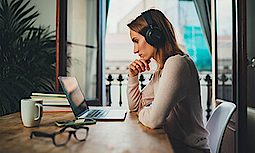 Eine junge Frau sitzt an einem Tisch und betrachtet nachdenklich ihren Laptop. Sie trägt ein schwarzes, kabelloses Headset. Im Hintergrund ist eine Pflanze und eine geöffnete Balkon-Tür. Auf dem Holztisch liegen Bücher, eine Tasse, ein Handy und eine Brille.