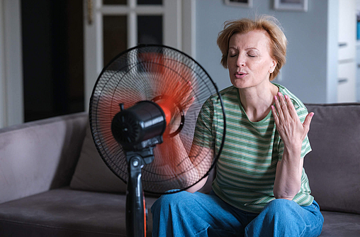 Frau im mittleren Altern sitzt vor einem Ventilator, ihr ist heiß.