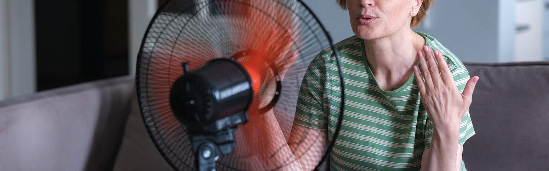 Frau im mittleren Altern sitzt vor einem Ventilator, ihr ist heiß.