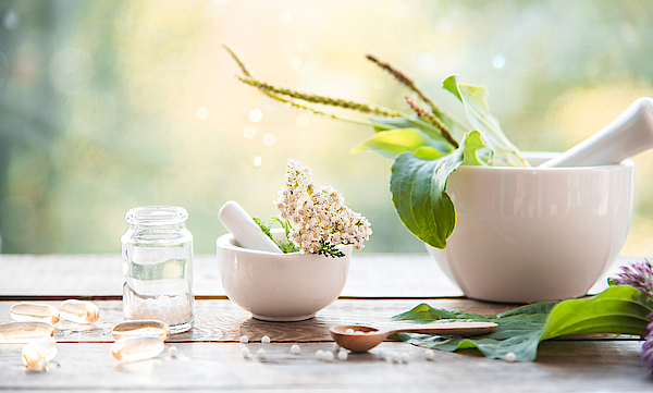 Auf einem Tisch steht ein Mörser mit Stößel. Drum herum sind verschiedene Pflanzen angeordnet. Sie alle wirken im Rahmen der Naturmedizin. Ein kleines Fläschen steht daneben. In dieses soll die Medizin eingefüllt werden. Es ist noch leer.