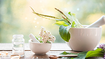 Auf einem Tisch steht ein Mörser mit Stößel. Drum herum sind verschiedene Pflanzen angeordnet. Sie alle wirken im Rahmen der Naturmedizin. Ein kleines Fläschen steht daneben. In dieses soll die Medizin eingefüllt werden. Es ist noch leer.
