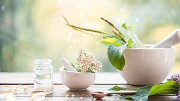 Auf einem Tisch steht ein Mörser mit Stößel. Drum herum sind verschiedene Pflanzen angeordnet. Sie alle wirken im Rahmen der Naturmedizin. Ein kleines Fläschen steht daneben. In dieses soll die Medizin eingefüllt werden. Es ist noch leer.