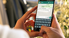 Nahaufnahme eines Handys, auf dem ein WhatsApp Chat geöffnet ist. Das Handy wird von zwei Händen gehalten.