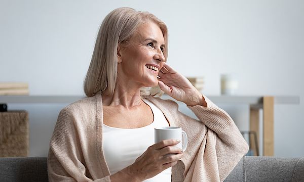 Eine ältere Frau sitzt auf dem Sofa. Sie hat graue Haare. In der Hand hält sie eine Kaffeetasse. Sie lacht und blickt in die Ferne.