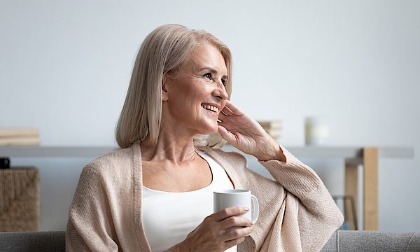 Eine ältere Frau sitzt auf dem Sofa. Sie hat graue Haare. In der Hand hält sie eine Kaffeetasse. Sie lacht und blickt in die Ferne.