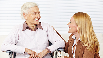 Eine ältere Dame sitzt lächelnd im Rollstuhl, sie unterhält sich mit einer jungen Frau.
