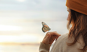 Auf der Hand einer Frau sitzt ein orange-schwarzer Schmetterling. Sie trägt eine orangene Mütze. Sie ist von hinten zu sehen.
