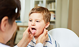 Einem kleinen Jungen wird beim Arzt der Hals abgetastet. Er streckt dabei die Zunge raus.