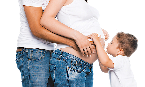 Eine schwangere Frau wird von hinten von ihrem Partner umarmt. Ein kleiner Junge küsst den schwangerschafts Bauch der Frau.
