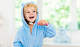 Blonder Junge in blauem Bademantel putzt lachend seine Zähne