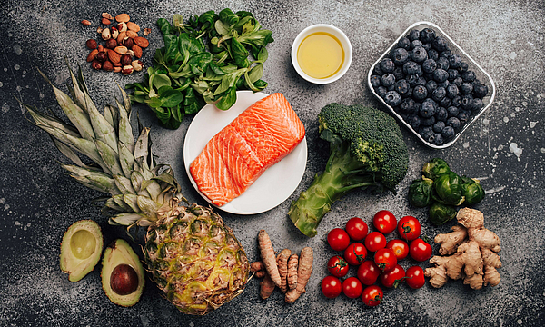 Ananas, Avocada, Oliven, Lachs, Brokolie und Ingver liegen auf einem dunklen Untergrund, sie gehören zu den entzündungshemmende Lebensmitteln.